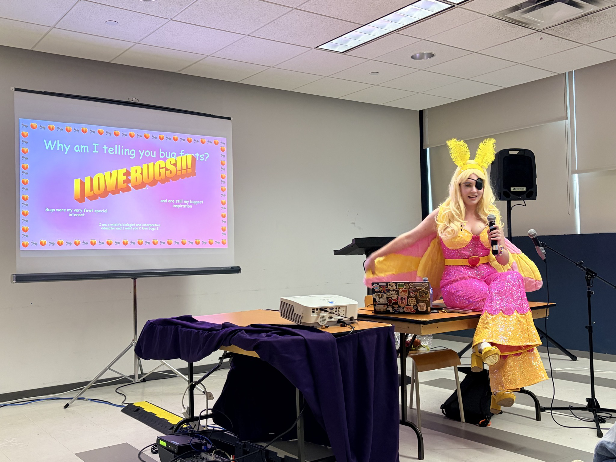 A photo of a woman dressed up as a rosy maple moth with a pink and yellow sequined costume, pink and yellow moth wigs, blond hair, yellow feather antennae, and an eyepatch sitting on a table holding a microphone while giving a lecture. On the screen in the back, there is a slideshow that says "I LOVE BUGS" in large text.